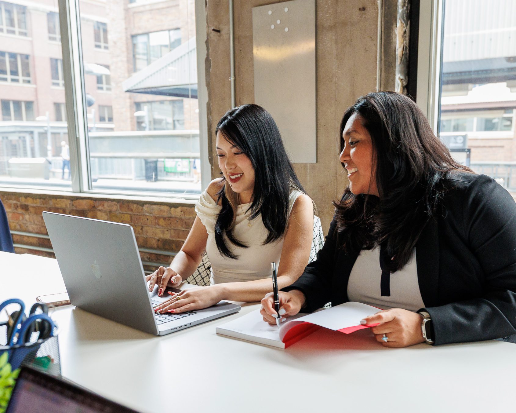 Two businesswomen sitting at a table in an office, looking at a laptop while smiling and taking notes collaboratively.