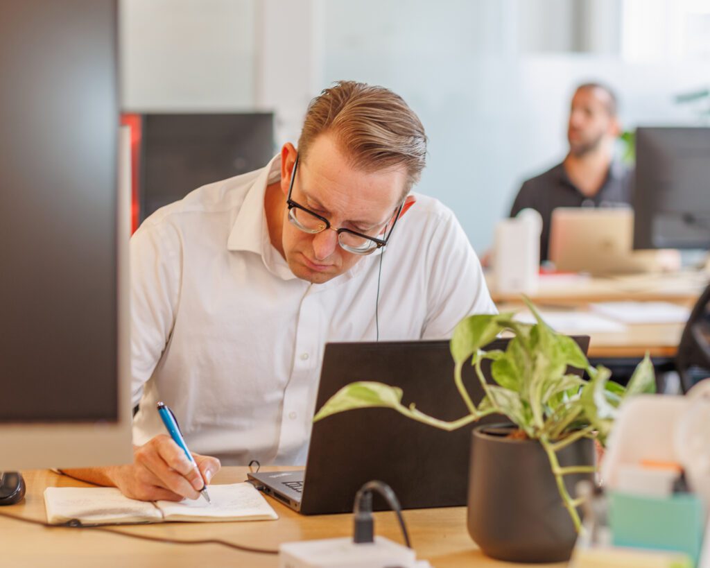 A man sitting at a desk with a laptop open, writing something in a notebook.