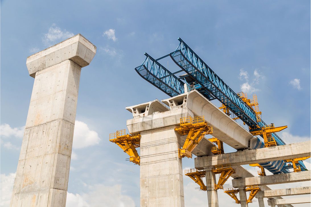 Concrete bridge under construction against a clear blue sky. Steel beams and yellow scaffolding highlight the engineering progress and industrial scale.